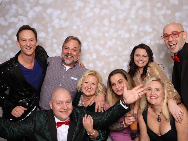 A group of adults smiling and posing together in a photo booth at a holiday party in Virginia Beach, standing in front of a bokeh backdrop and wearing festive attire.