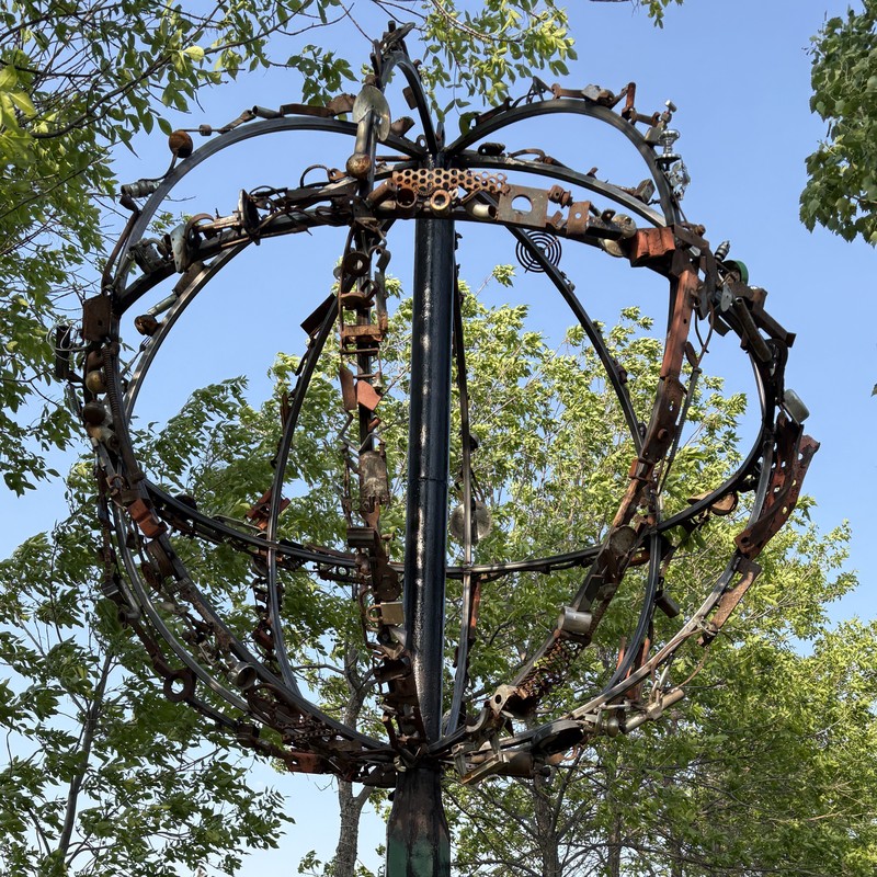 Monumental 6-foot spherical metal sculpture titled 'Space Debris,' handcrafted by Saskatoon artist Rik Luczka from recycled steel scraps and found objects. Mounted atop a tall industrial pole, this celestial-inspired globe sculpture features integrated night lighting, creating a glowing abstract silhouette. A landmark piece of Saskatchewan public art, it showcases expert welding and large-scale metal fabrication. Available for custom commercial or municipal art commissions in Saskatoon.