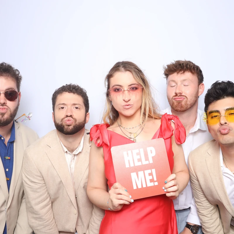 A group of wedding guests poses in front of a white photo booth backdrop, with one guest holding a red “HELP ME!” sign. Captured by VA Shoreline Events using a luxury DSLR photo booth at a Virginia Beach wedding.