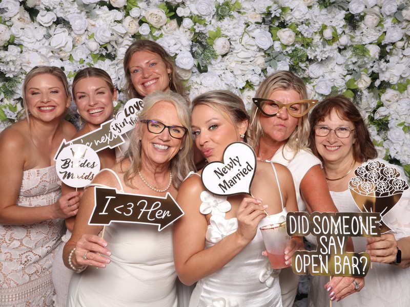 Group of women posing together in a wedding photo booth in front of a white textured floral wall backdrop, holding wedding-themed props and celebrating the newly married couple.