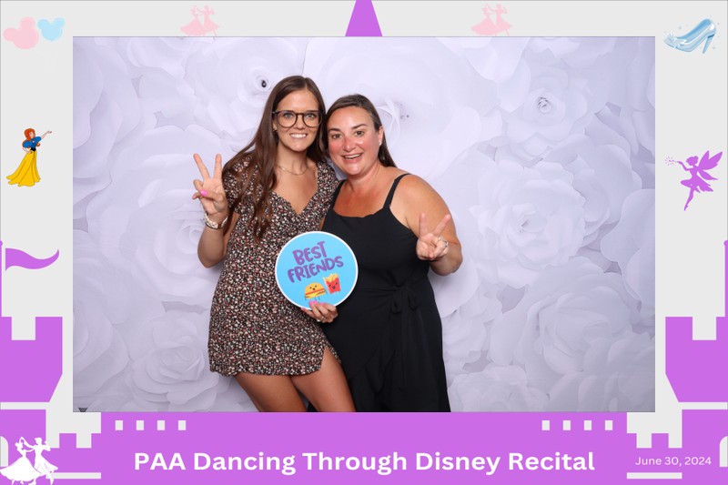 Two friends posing together in a photo booth in front of a white floral backdrop, holding a “Best Friends” sign and making peace signs during the PAA Dancing Through Disney recital on June 30, 2024.