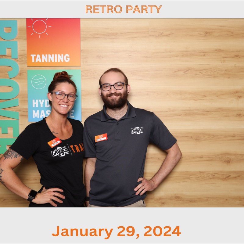 Two gym staff members are standing side by side and smiling in front of a wood-panel backdrop with colorful signage for tanning and hydromassage services. The date “January 29, 2024” is displayed below the photo.