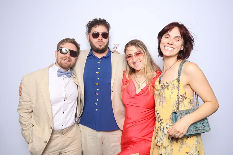 Four wedding guests smiling and posing together in a photo booth at a Virginia Beach celebration. They are wearing bright, stylish outfits and sunglasses against a clean, light backdrop.