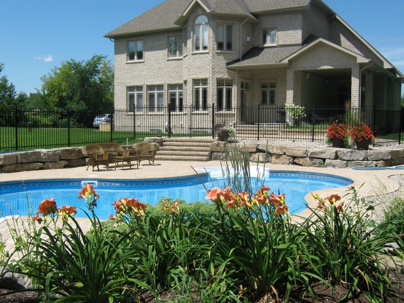 A beautifully designed backyard pool with curved stone borders, a spacious patio area, and lush flowerbeds in front of a large stone home. The design highlights Stonebuilders’ craftsmanship in creating inviting outdoor living spaces.