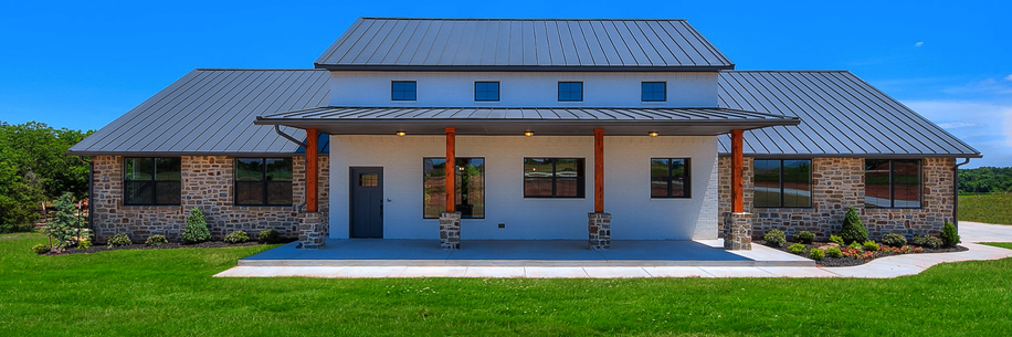 The Snider Custom Home showcases a modern farmhouse exterior with a gray metal roof, white siding, and stone details. Three wooden porch columns frame the entryway, surrounded by landscaped greenery and an open lawn. Built by Merryfield Homes in Mustang, Oklahoma, the home reflects craftsmanship and rural elegance.