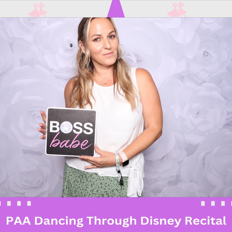 A woman is smiling and holding a sign that says “Boss Babe” while standing in front of a white floral backdrop. The photo frame features a Disney-inspired theme with castle silhouettes and small character illustrations. The text at the bottom reads “PAA Dancing Through Disney Recital” with the date June 30, 2024.