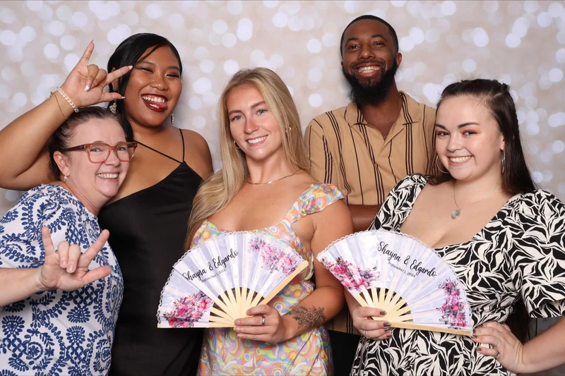A group of five wedding guests smiling and posing together in a photo booth in Virginia Beach. They are holding decorative floral hand fans and standing in front of a soft bokeh backdrop.