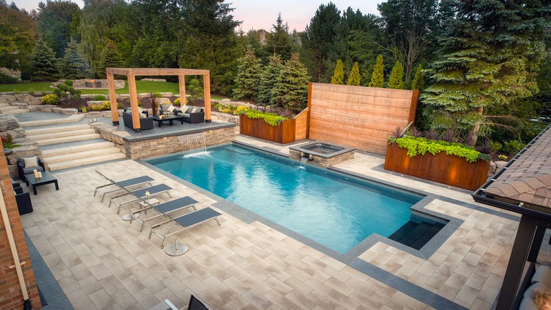 A luxury backyard oasis with a rectangular pool surrounded by light stone pavers, integrated water features, and a raised lounge area beneath a wooden pergola. Custom planters and layered stonework add warmth and sophistication, showcasing Stonebuilders’ mastery in pool and patio design.