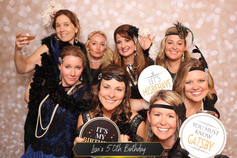 Group of women dressed in vintage-inspired attire posing together in a photo booth in front of a champagne sparkle backdrop, holding playful signs during Lisa’s 50th birthday celebration.