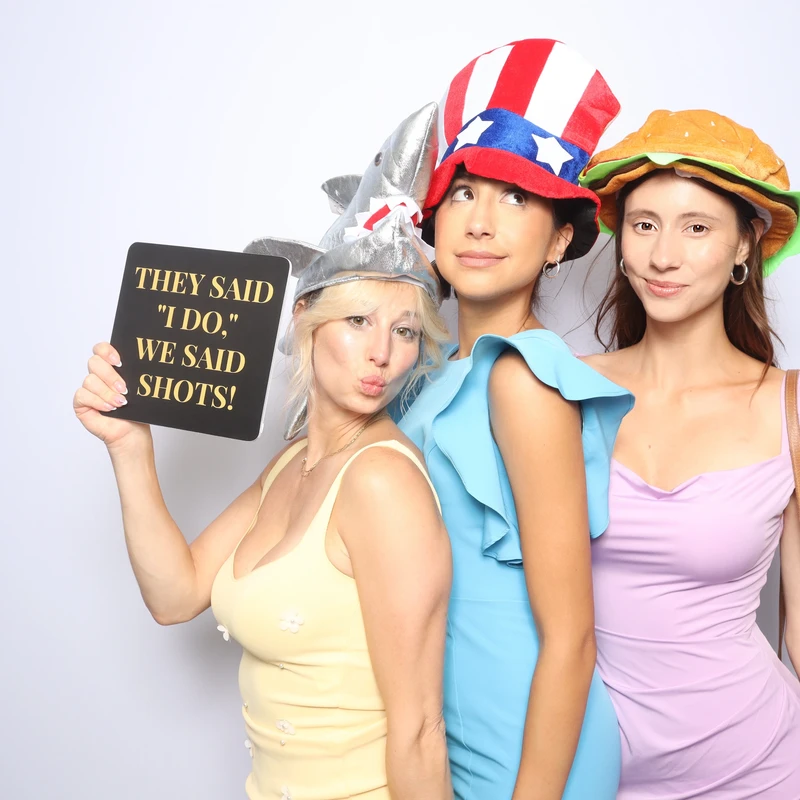 Three wedding guests pose in front of a white photo booth backdrop wearing fun hats and holding a “They said I do, we said shots!” sign. Captured by VA Shoreline Events at a Virginia Beach wedding, featuring the luxury DSLR photo booth setup.
