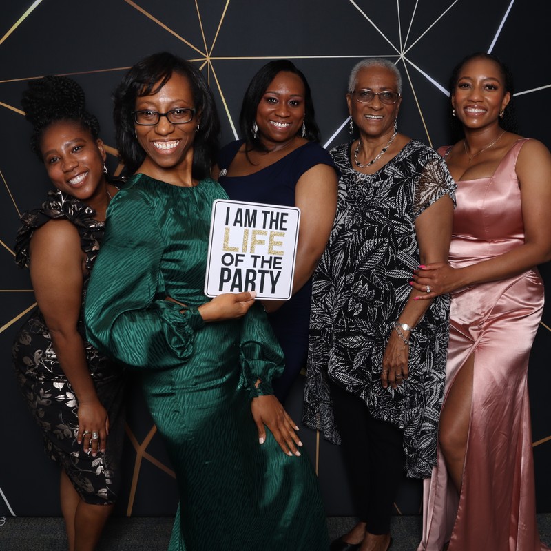 Group of women posing together in a photo booth in front of a modern black backdrop with gold geometric lines, holding a fun “I am the life of the party” sign during a birthday celebration.