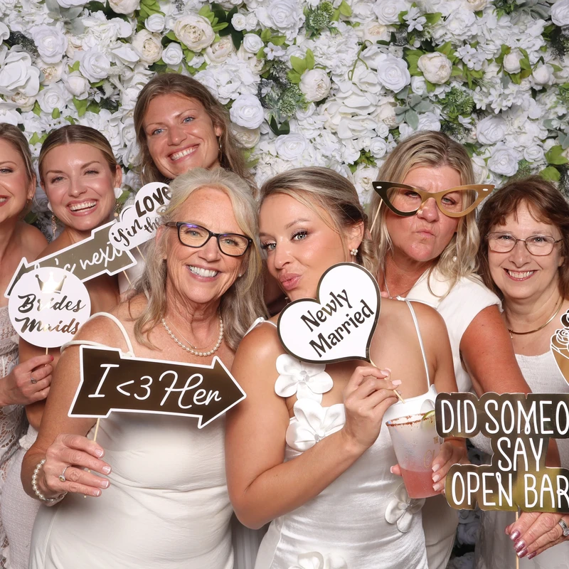 A group of bridesmaids and wedding guests pose in front of a white floral backdrop inside the VA Shoreline Events luxury DSLR photo booth. Captured at a Virginia Beach wedding with fun props and instant sharing.