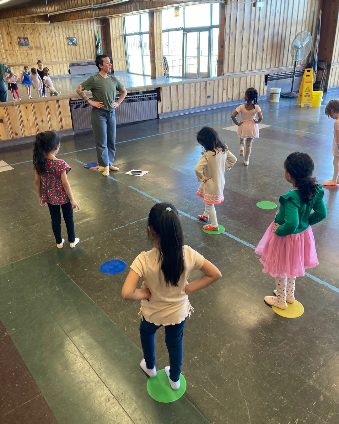 Kait leads beginning ballet students. They stand on floor dots in first position with their hands on their hips
