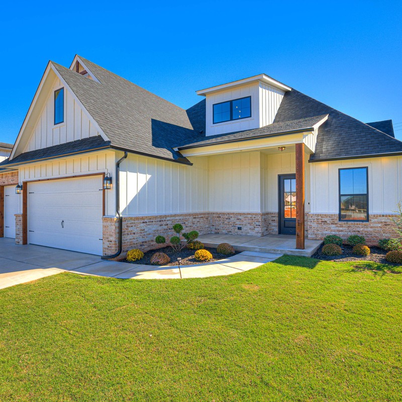 Ground-level front angle view highlighting the farmhouse exterior, garage doors, and landscaped front yard of 10408 SW 51st Street in Mustang Oklahoma.