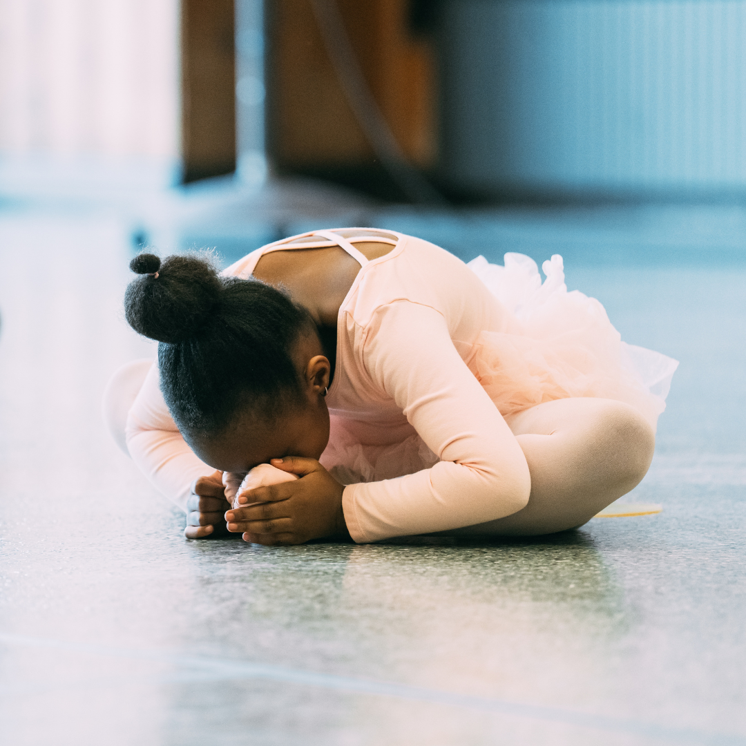 A ballet student sits in the butterfly position, folding over, touching their face to their toes.