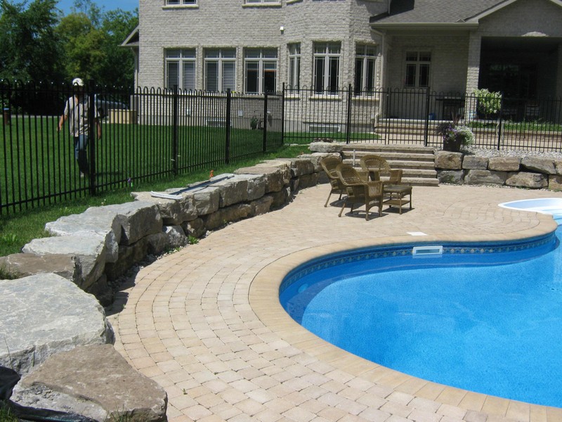 A sophisticated backyard featuring a custom pool surrounded by light-toned interlock paving and a natural stone retaining wall. The design includes steps leading to the home and a seating area, showcasing Stonebuilders’ expertise in combining natural and modern hardscaping elements.
