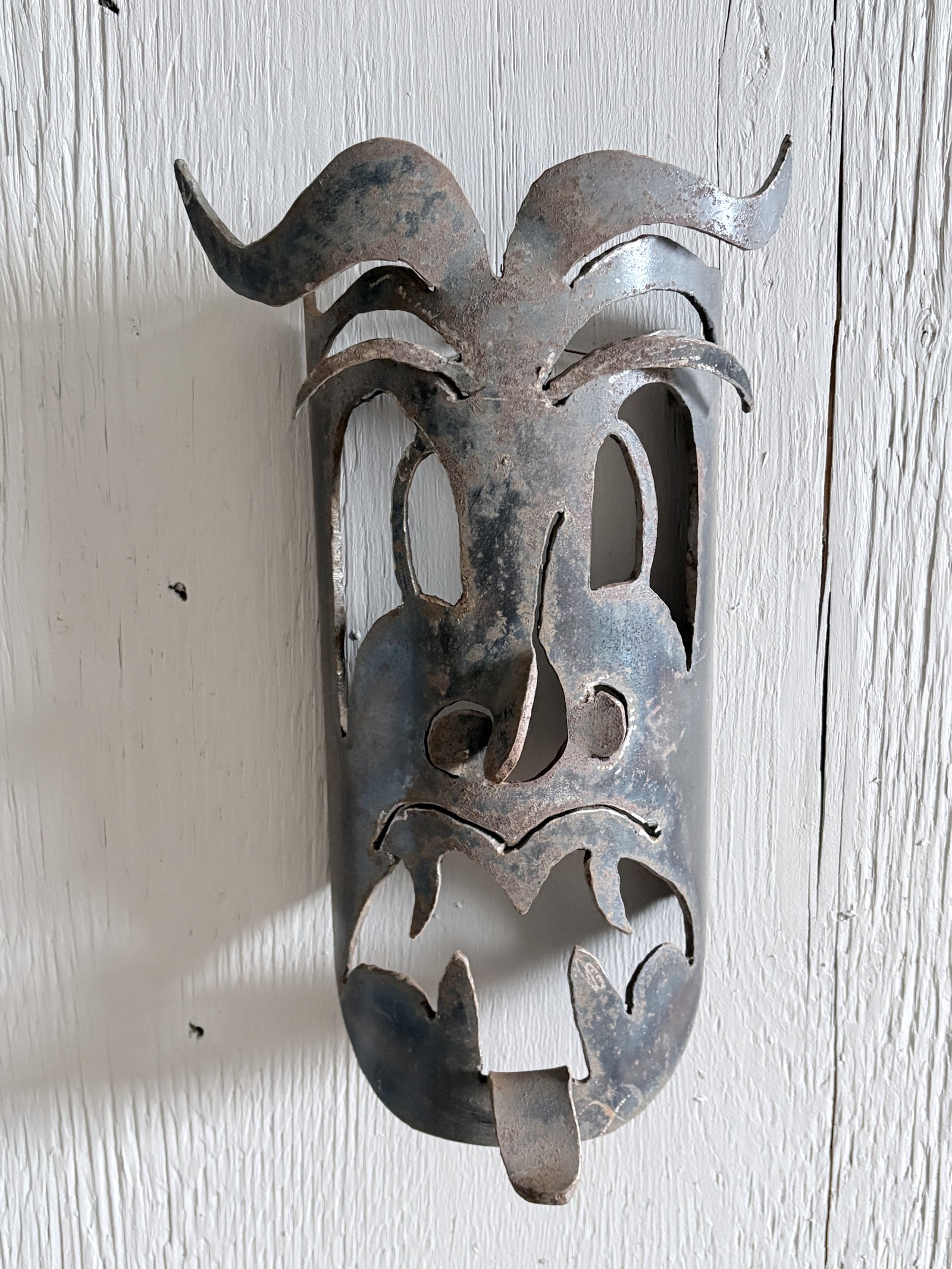 Unique recycled steel mask titled 'Bad Dream' by Saskatoon artist Rik (Rick Luczka). This expressive wall-mounted sculpture features exaggerated facial details, including a prominent curved nose, bushy metal eyebrows, and a tongue sticking out. Mounted on a textured white wood base, this one-of-a-kind piece of Saskatchewan metal art adds a bold, character-driven touch to modern home decor.