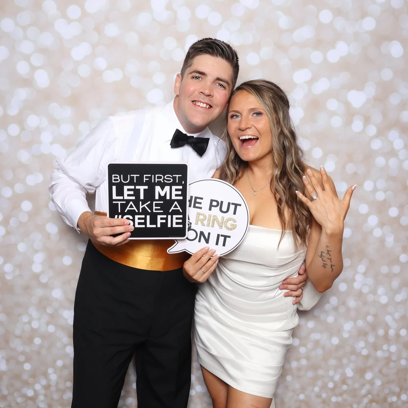 A newly married couple poses in front of a gold bokeh backdrop holding signs that read ‘But first, let me take a selfie’ and ‘He put a ring on it.’ Captured by VA Shoreline Events’ luxury DSLR photo booth at a Virginia Beach wedding.