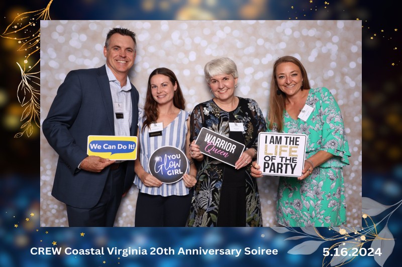 Four guests smiling and posing together in a photo booth at the CREW Coastal Virginia 20th Anniversary Soiree on May 16, 2024, holding celebratory signs in front of a soft bokeh-style backdrop.