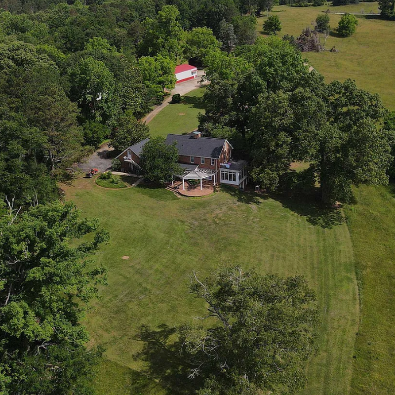 Aerial view of the 1978 brick home and the 100x32 multipurpose building with red roof, surrounded by mature trees