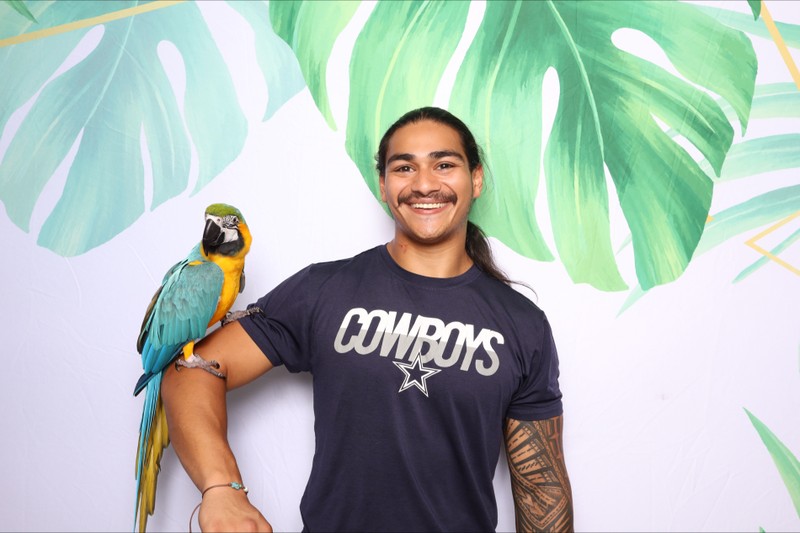 A smiling guest posing in a photo booth with a vibrant parrot perched on his arm, standing in front of a tropical palm leaf backdrop at Landon’s pool party fundraiser.