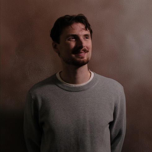 A person with dark hair and short facial hair wearing a light gray sweater, standing against a textured brown wall and looking slightly upward with a gentle smile.