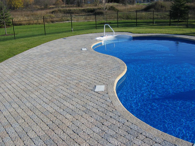 An elegant outdoor pool design featuring gently curved stone pavers forming a seamless border around crystal-blue water. The open lawn and iron fencing provide a polished, spacious feel—showcasing Stonebuilders’ craftsmanship in modern pool surrounds.