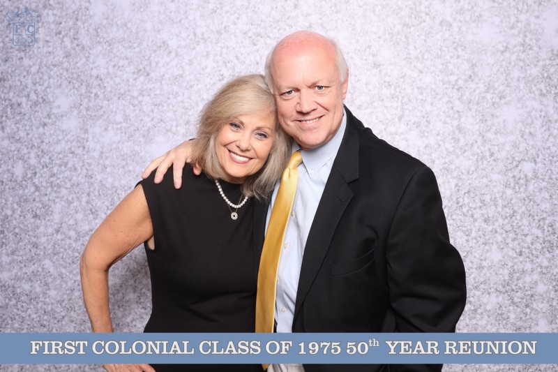 A couple smiling and standing together in front of a light textured photo booth backdrop during the First Colonial High School Class of 1975 50th year reunion celebration.