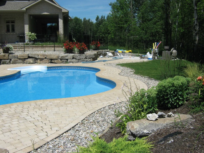 A bright backyard scene featuring a clear blue pool surrounded by light-colored interlock paving and decorative stone edging. Lush greenery and a natural stone retaining wall complement the clean, modern design crafted by Stonebuilders Landscaping.