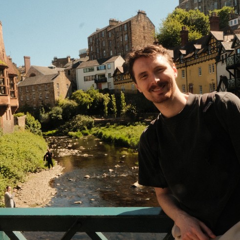 A person standing in Dean Village, Edinburgh, with historic stone buildings and the Water of Leith visible in the background.