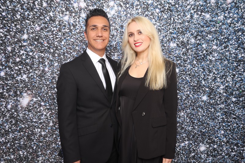 A well-dressed couple smiling in front of a silver glitter photo booth backdrop at a holiday party, captured by a professional DSLR photo booth setup.