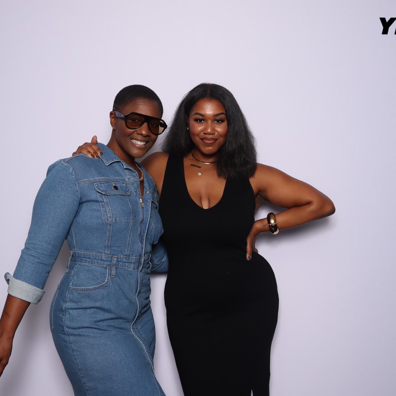 Two women are standing together and smiling in front of a plain light backdrop. One is wearing a denim jumpsuit and sunglasses, and the other is wearing a black sleeveless dress. The word “YELLOW” appears in the upper right corner of the image.