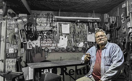Saskatoon metal artist Rick Luczka (Rik) at his workbench in his creative studio. The workshop features a collection of tools, vintage signs, and license plates, showcasing the authentic environment where he handcrafts recycled steel sculptures and custom metal art in Saskatchewan.