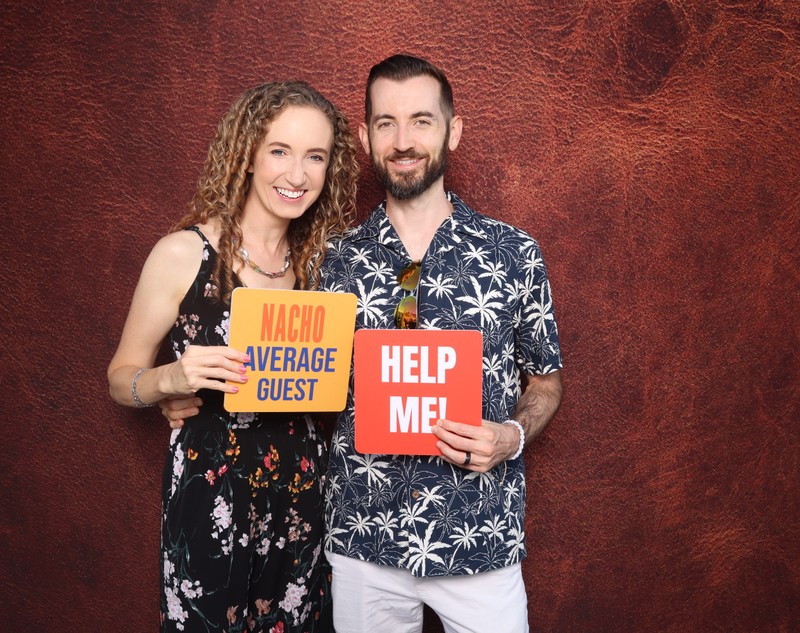 A smiling couple posing together in a photo booth in front of a warm textured backdrop, holding playful signs during a birthday celebration photo booth experience.