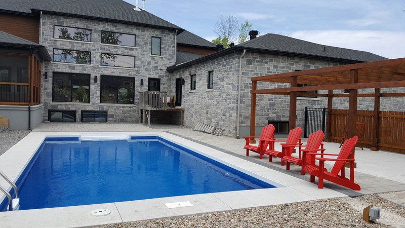 A clean and contemporary backyard pool area featuring a rectangular blue pool bordered by light concrete and interlock paving. Red lounge chairs sit beneath a wooden pergola beside a gray stone home, highlighting Stonebuilders’ craftsmanship in modern pool surround design.