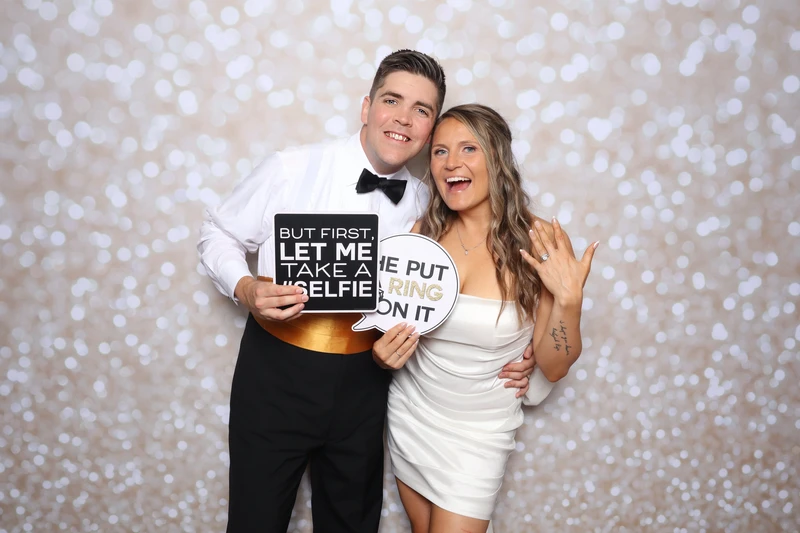 A newlywed couple smiling and posing in a photo booth in Virginia Beach. They are holding playful wedding-themed signs and standing in front of a soft bokeh backdrop.