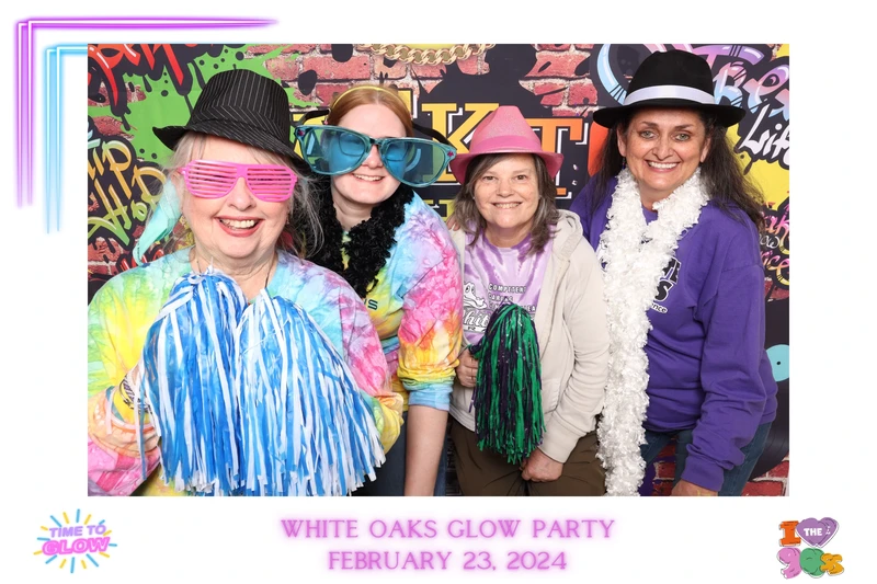 Four women smiling and posing in a photo booth at a glow-themed party in Virginia Beach, wearing colorful tie-dye shirts, hats, oversized sunglasses, and fun props in front of a vibrant graffiti-style backdrop.