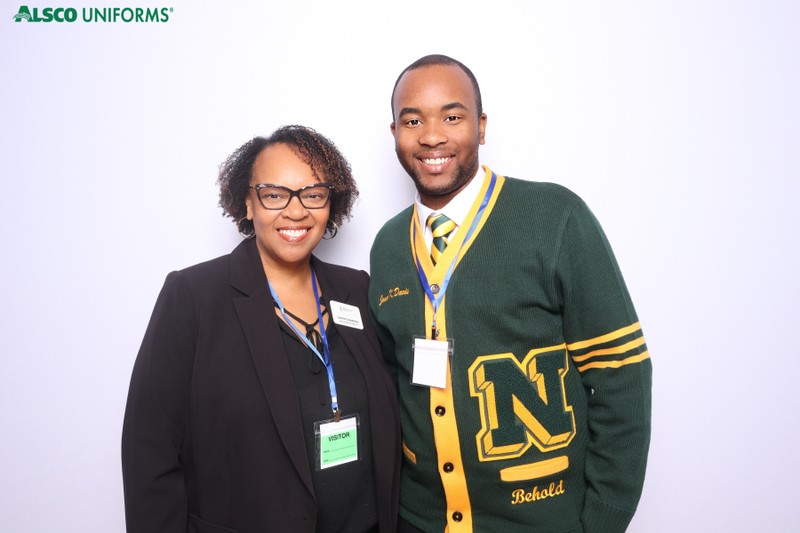 Two colleagues smiling and posing together in a photo booth in front of a clean white backdrop during an Alsco Uniforms corporate event, wearing professional attire and event badges.