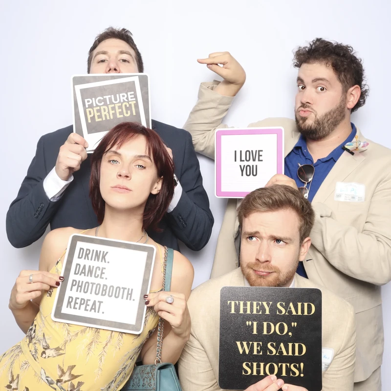 Four guests posing together in a photo booth at a wedding in Virginia Beach. Each person is holding a sign: “Picture Perfect,” “I Love You,” “Drink. Dance. Photobooth. Repeat.,” and “They said ‘I do,’ we said shots!” Perfect for weddings and celebrations across Virginia Beach, Norfolk, and Chesapeake.