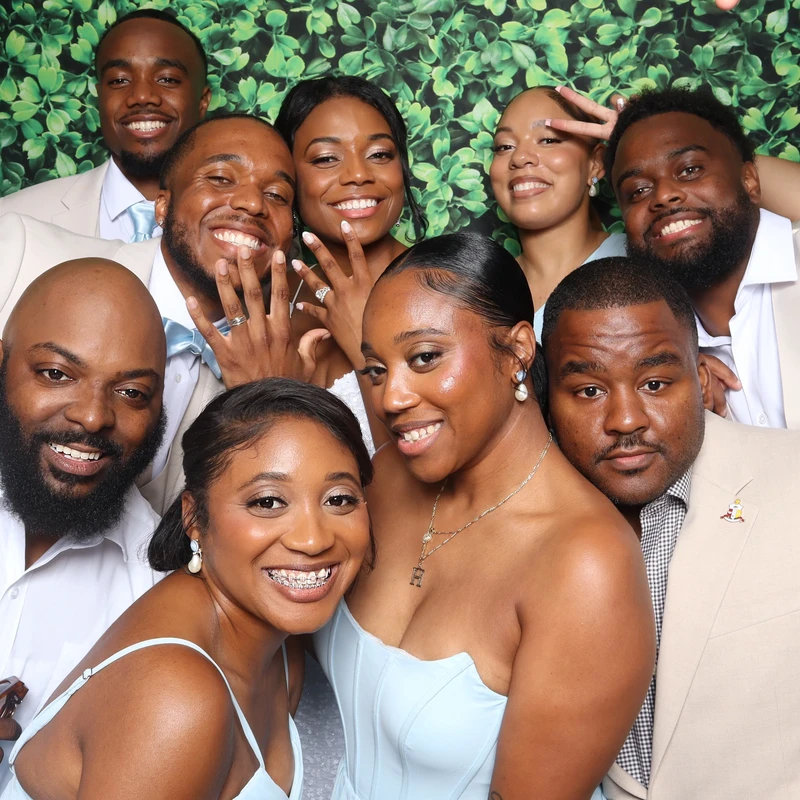A group of bridesmaids and wedding guests pose in front of a white floral backdrop inside the VA Shoreline Events luxury DSLR photo booth. Captured at a Virginia Beach wedding with fun props and instant sharing.