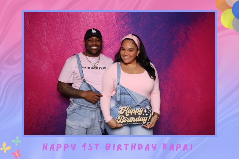 A smiling couple posing together in a photo booth in front of a vibrant pink textured backdrop, holding a “Happy Birthday” sign during Kapri’s first birthday celebration.