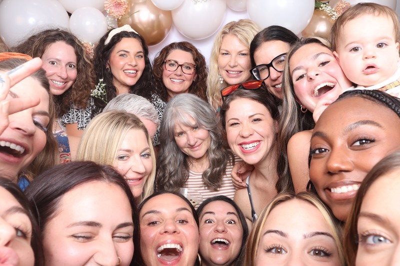Large group of guests smiling and posing closely together in a photo booth with a white and gold balloon backdrop, creating a fun and energetic celebration photo.