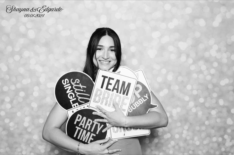 Black-and-white photo booth portrait of a smiling guest holding wedding-themed props in front of a soft sparkle backdrop, taken at Shayna and Edgardo’s wedding on September 1, 2024.