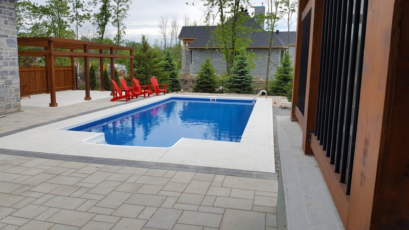 A contemporary pool design featuring a rectangular blue pool bordered by smooth concrete and interlock pavers. A wooden pergola with red lounge chairs adds warmth and contrast, showcasing Stonebuilders’ expertise in modern pool patio installations.