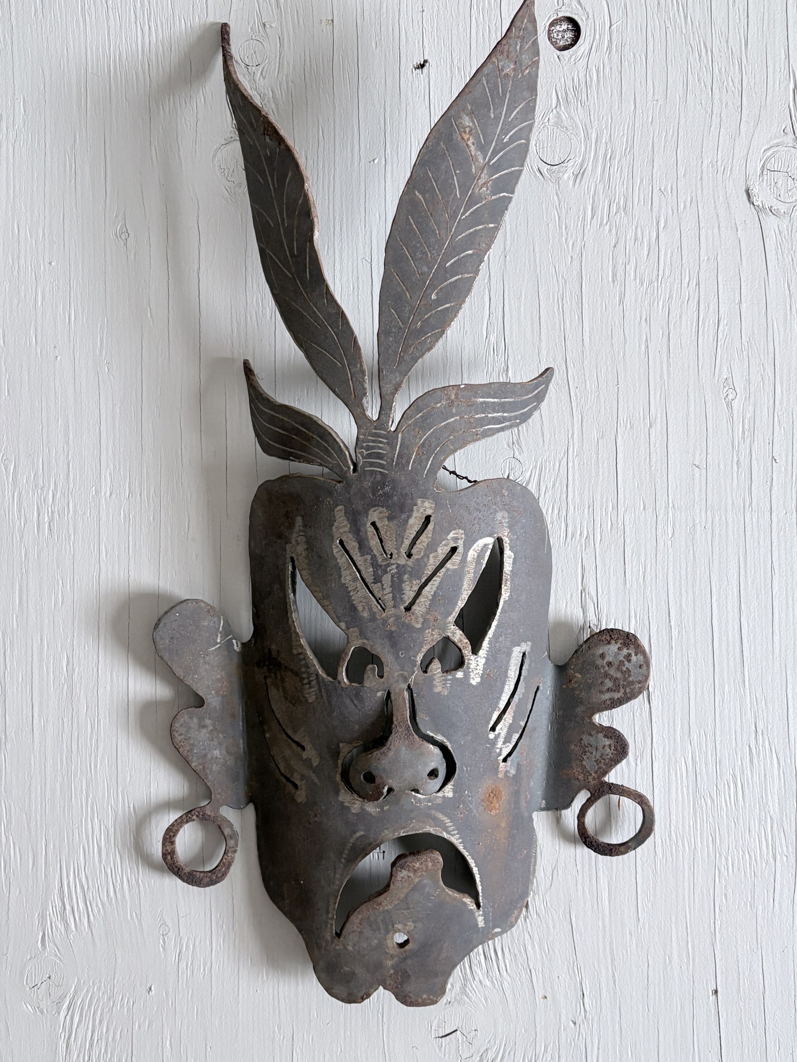 Handcrafted metal mask titled 'Medicine Man' by Saskatoon artist Rik (Rick Luczka). This expressive welded steel face features intricate engraved details, circular ear ornaments, and two large stylized leaf shapes rising from the head. Mounted on a weathered gray wood base, this unique wall-mounted metal sculpture is a standout piece of Saskatchewan-made fine art for home or office decor.