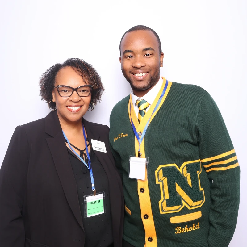 Two people smiling and standing together in front of a clean white backdrop at a professional event. Both are wearing business attire with visitor badges and lanyards, posing for a polished photo booth portrait. The image conveys a friendly and professional atmosphere suitable for corporate networking and university or organizational events.