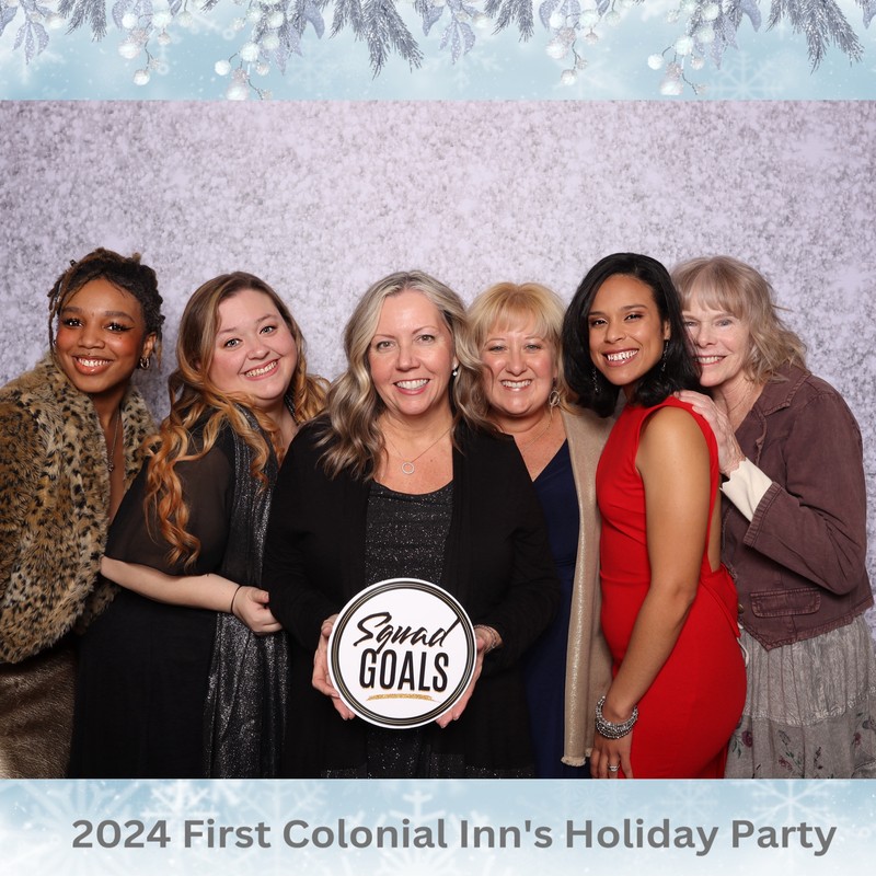 Six women are standing close together and smiling in front of a snowy textured backdrop. They are dressed in winter party attire, and one is holding a circular sign that says “Squad Goals.” The photo has a light blue winter border with snowflakes and reads “2024 First Colonial Inn’s Holiday Party” at the bottom.