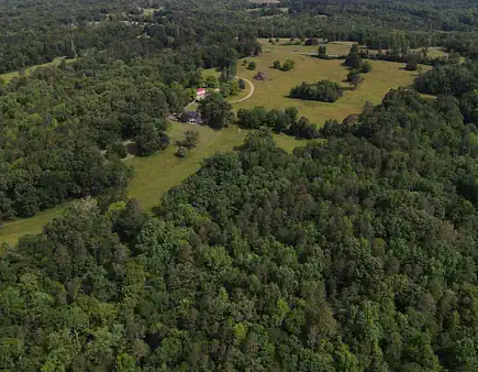 Aerial view of a large wooded property with dense green forest surrounding open grassy fields, winding dirt paths, and a small building nestled among the trees.”