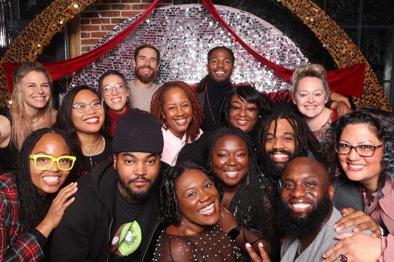 A large group of smiling guests posing closely together at the Yellow Holiday Social 2025 in front of a silver sequin photo booth backdrop with red holiday draping and festive décor.