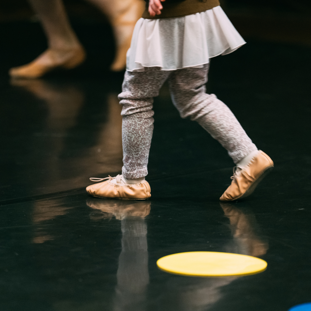 A close-up of a ballet student in grey leggings and ballet slippers. A yellow floor dot on black marley.